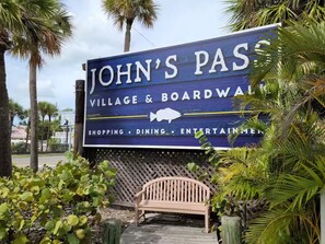 Schild für 'JOHN'S PASS VILLAGE & BOARDWALK' mit aufgelisteten Annehmlichkeiten wie Einkaufsmöglichkeiten, Gastronomie und Unterhaltung.