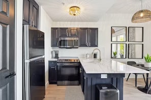 A modern kitchen with dark cabinetry, stainless steel appliances, a central island with a sink, and a pendant light fixture. - Forever Sunshine - Fully Renovated (St. Petersburg)