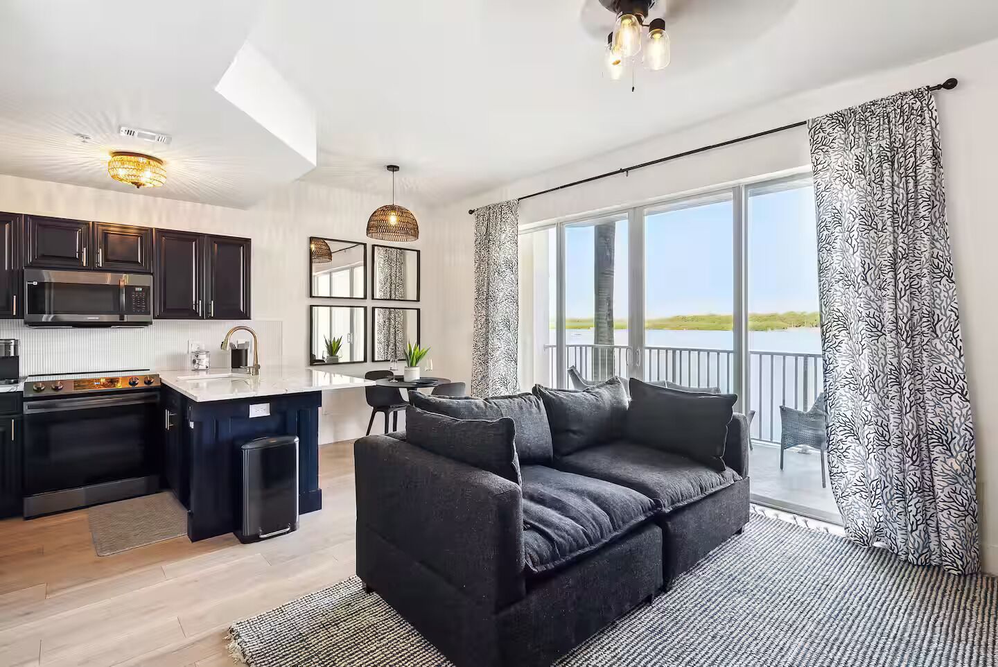 Eine moderne Küche mit dunklen Schränken, ein Essbereich mit grauem Sofa und große Fenster mit Blick ins Freie.