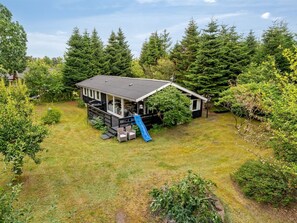 Eine Holzhütte mit Terrasse, Rutsche und umzäunter Garten, umgeben von Bäumen.