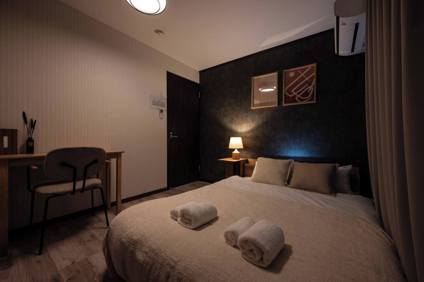 A hotel room with a bed, a desk, a chair, a lamp, and a wall-mounted air conditioning unit.
