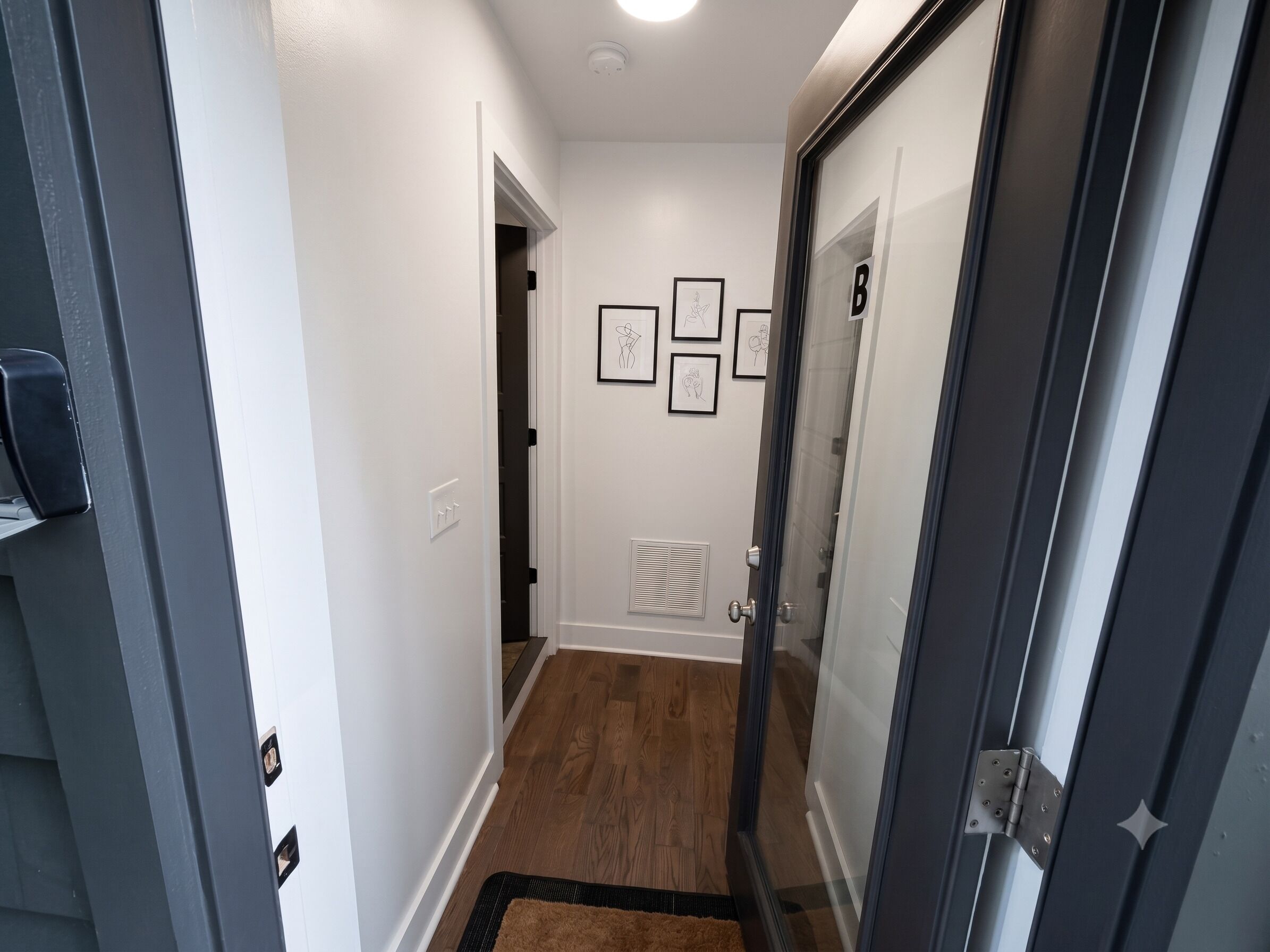 Un couloir avec une porte sur la droite, une grille d’aération sur le mur et trois tableaux encadrés accrochés au mur.
