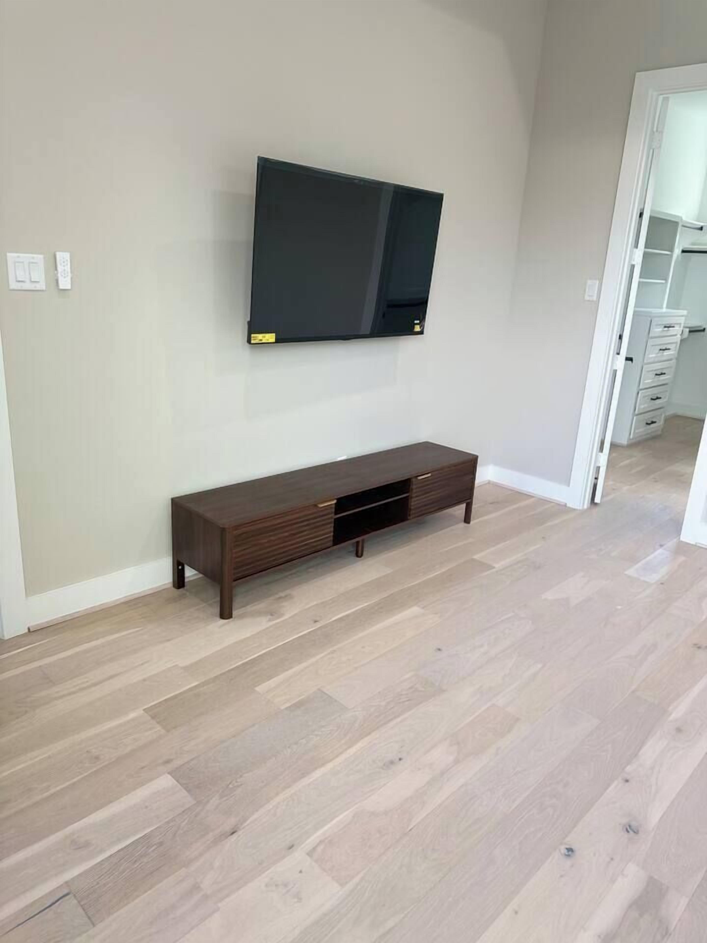 A room with a flat-screen TV mounted on the wall, a wooden console table, and a light-colored wooden floor.