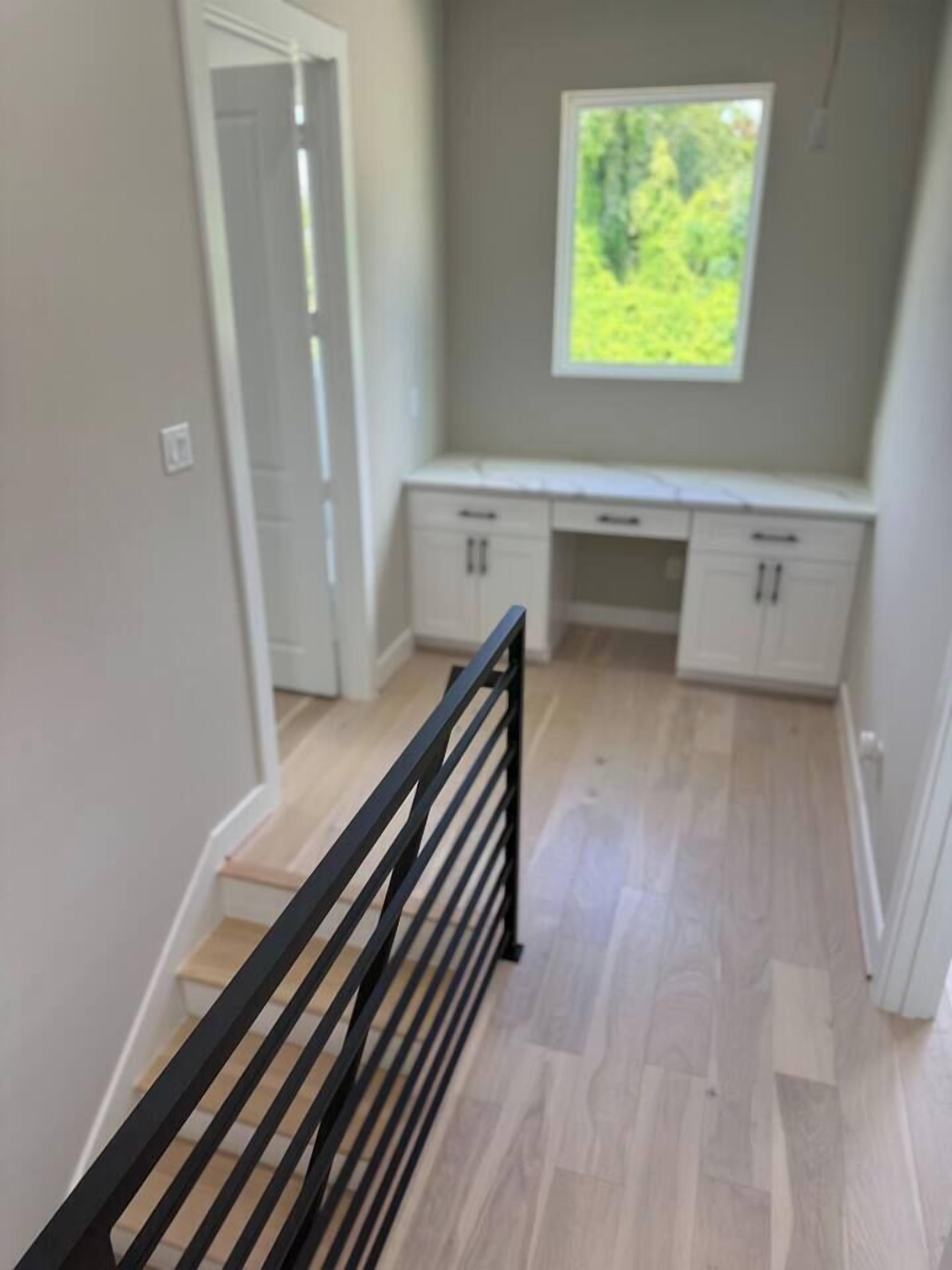 A narrow hallway with a wooden floor, a black railing, and a window with a view of greenery.