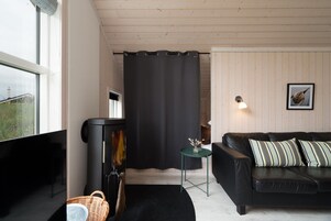 Ein gemütliches Wohnzimmer mit einem schwarzen Sofa, einem kleinen Beistelltisch, einem schwarzen Heizgerät und einem Fenster mit Blick auf Grünflächen.