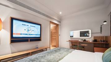 Una habitación de hotel moderna con una amplia televisión de pantalla plana montada en la pared, un banco de madera, una cama con un edredón texturizado, un escritorio con una lámpara y un espejo.