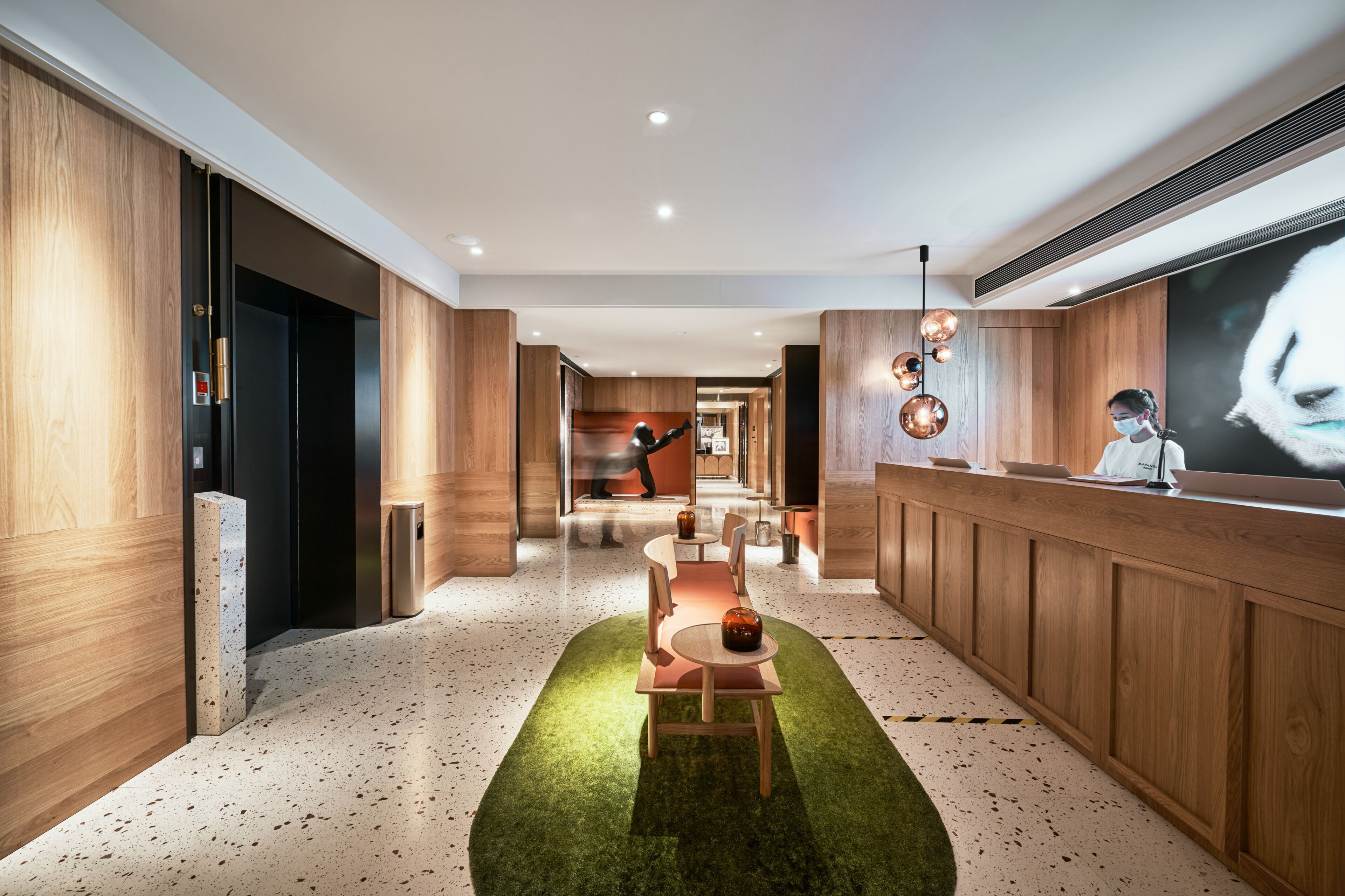 A modern hotel lobby with a reception desk, a seating area with a green rug, and wooden paneling on the walls.