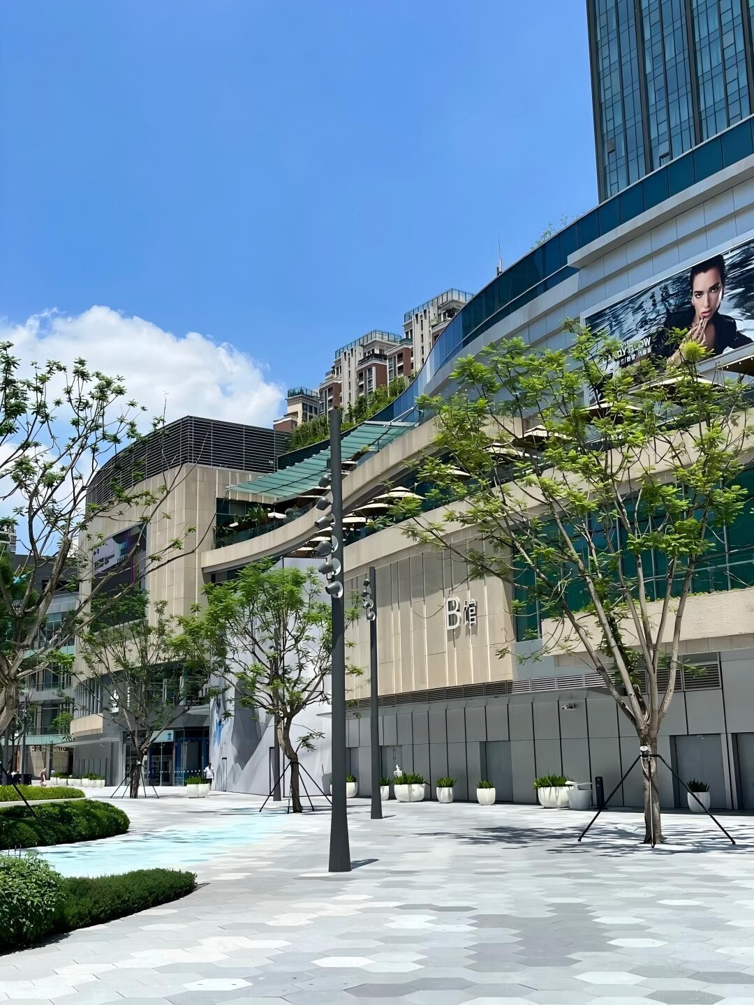 Un edificio moderno con un gran anuncio, mesas al aire libre y un área ajardinada con árboles.