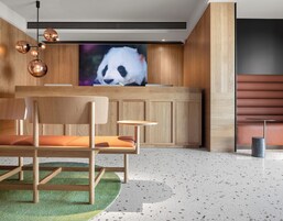 Un espace d’accueil moderne avec un comptoir de réception en bois, un canapé avec des coussins orange, une table ronde et une grande image de panda sur le mur.