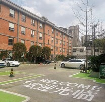 Un edificio de varios pisos con fachada de ladrillo, una zona de estacionamiento con automóviles y un espacio verde con árboles.