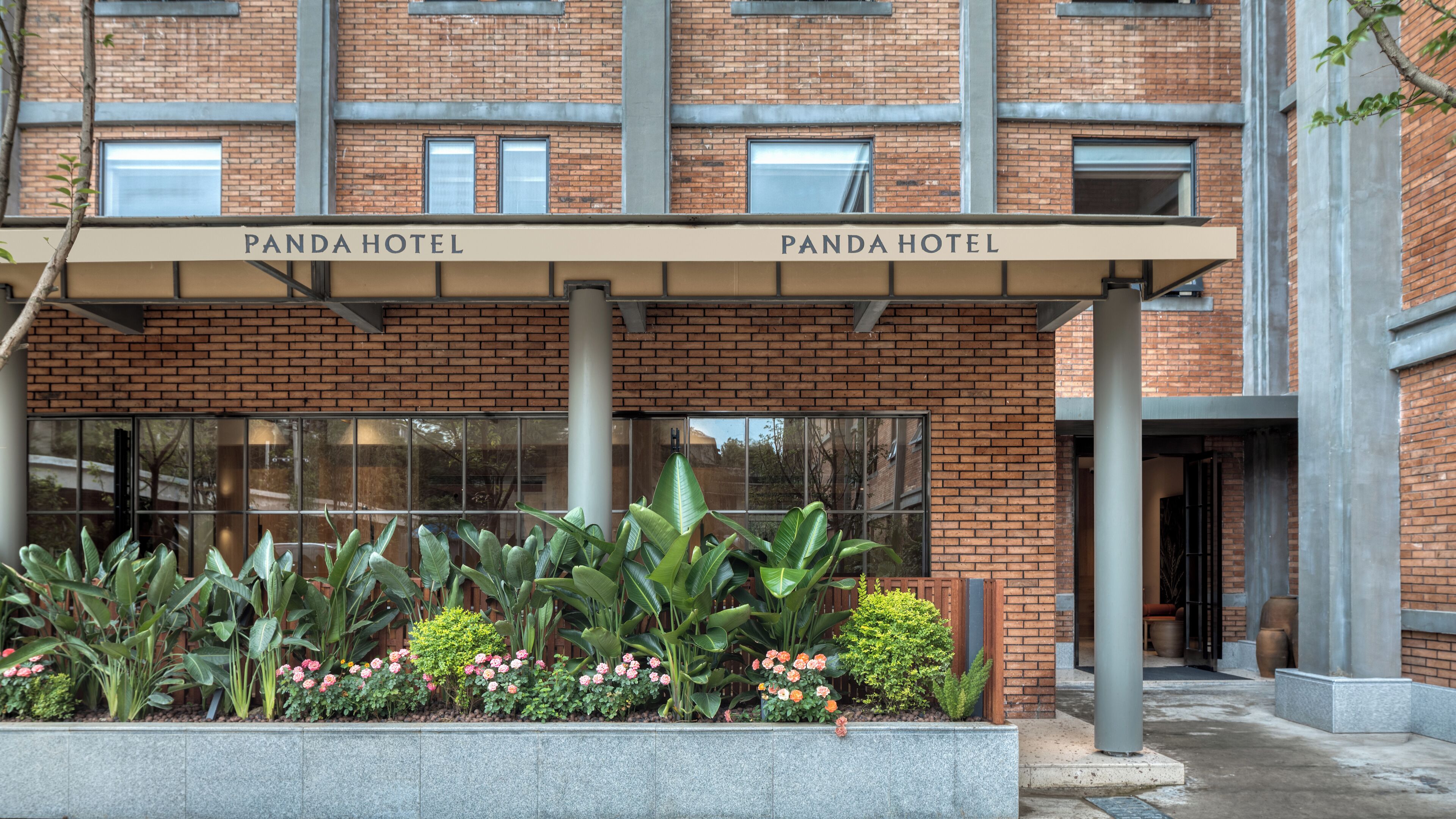 Entrada de un hotel con dos letreros que indican 'Panda Hotel' sobre la entrada. Hay una jardinera con diversas plantas y flores frente a la entrada.