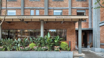 Entrada de un hotel con dos letreros que indican 'Panda Hotel' sobre la entrada. Hay una jardinera con diversas plantas y flores frente a la entrada.
