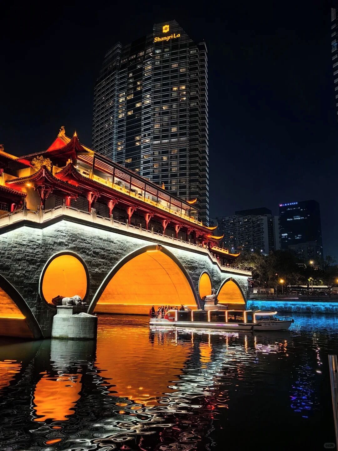 A traditional bridge with illuminated arches over a body of water, with modern buildings in the background.