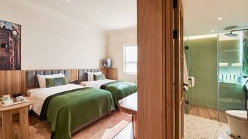 A hotel room with two beds, a bedside table, a nightstand, a window, and a shower area.