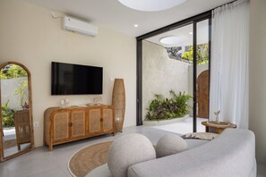 A modern living room with a flat-screen TV, a wooden cabinet, a large mirror, a sofa, and a view of a garden through a sliding glass door.