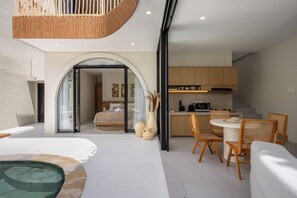 Una habitación de hotel moderna con un vidrio que da a un dormitorio, una zona de comedor con muebles de madera y una pequeña cocina.