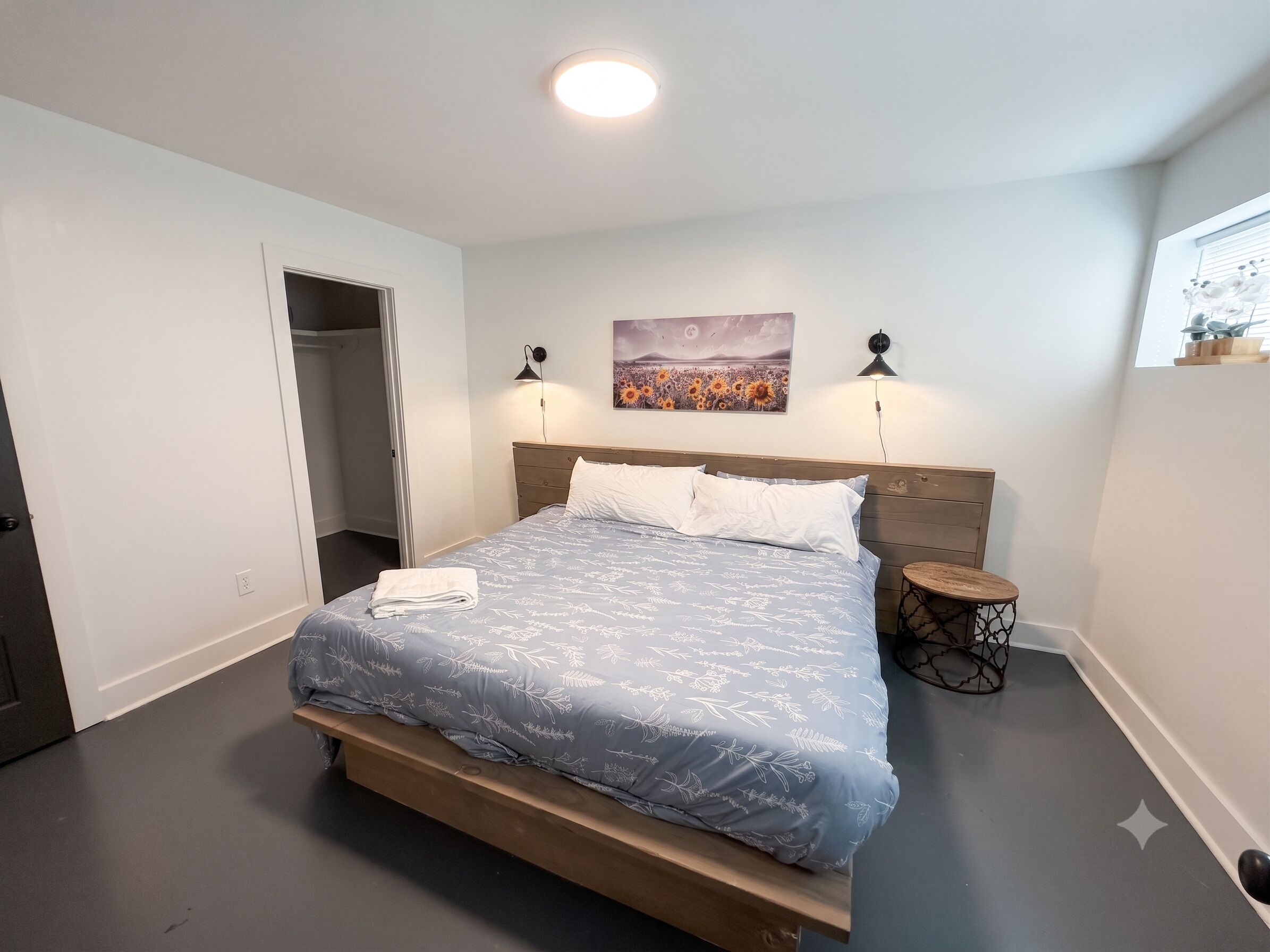Ein Schlafzimmer mit einem Bett, einem Nachttisch, einer Wandlampe, einem Gemälde und einem Fenster mit Jalousien.