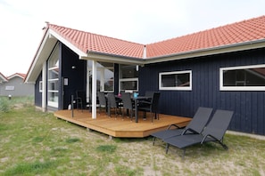Ein modernes Haus mit einer überdachten Terrasse, einem Essbereich mit Stühlen und einem Tisch sowie einem Deck mit Liegestühlen.