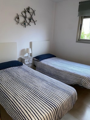 Een kamer met twee eenpersoonsbedden, een nachtkastje met een lamp, een decoratie aan de muur en een raam.