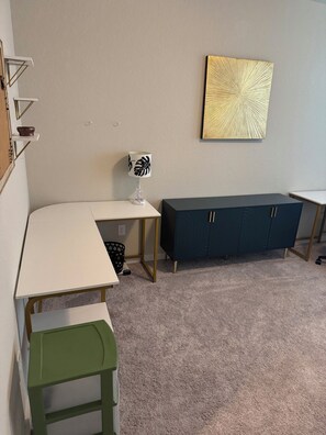 Ein Zimmer mit einem weißen Schreibtisch, einem grünen Stuhl, einem dunkelblauen Schrank und einem Wandbild.