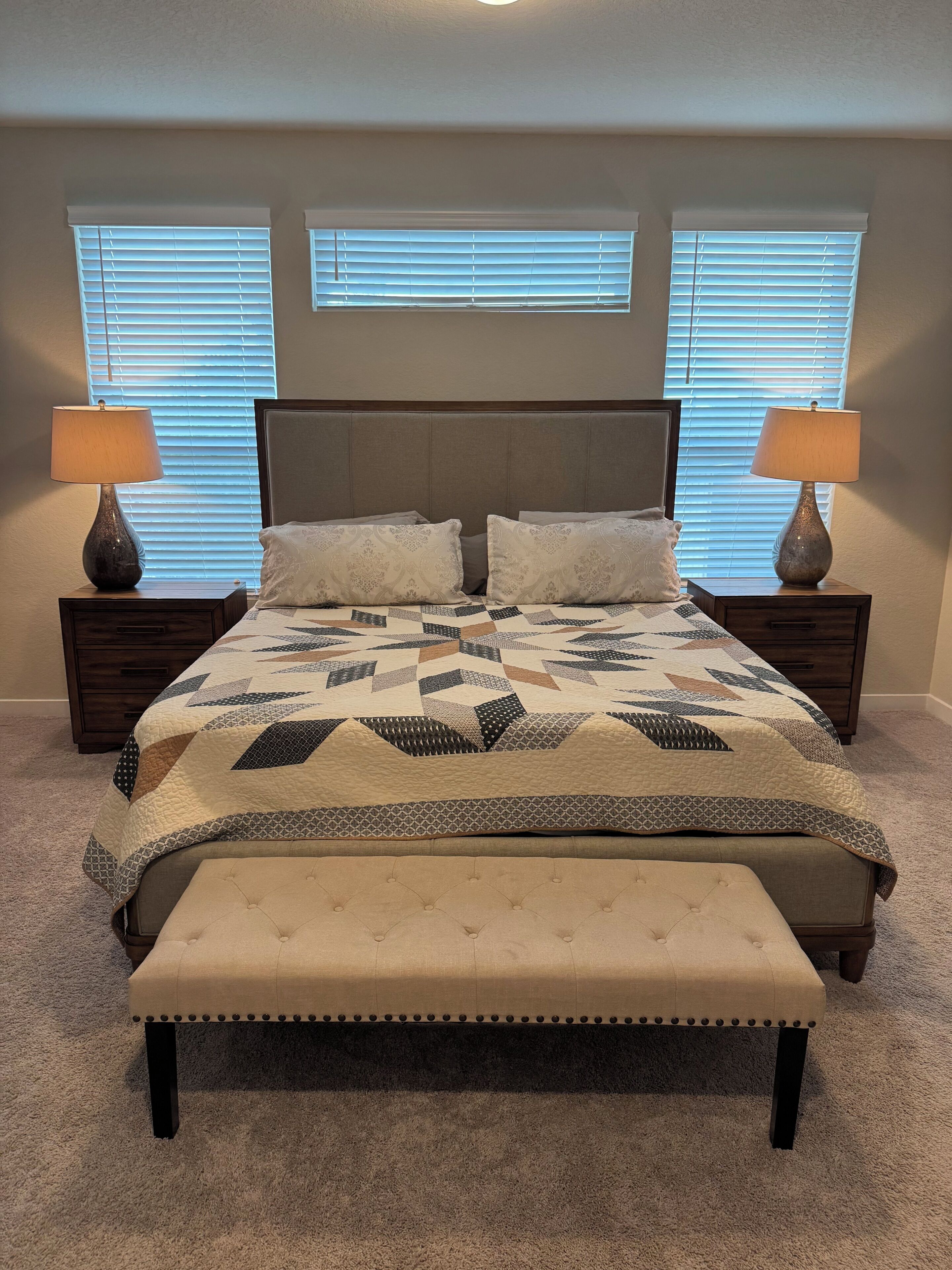 Ein Schlafzimmer mit einem Bett, zwei Nachttischen, einer Bank und drei Fenstern mit Jalousien.