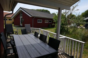 Ein überdachter Außenbereich mit Tisch und Stühlen, einem weißen Geländer und Blick auf ein rotes Haus im Hintergrund.