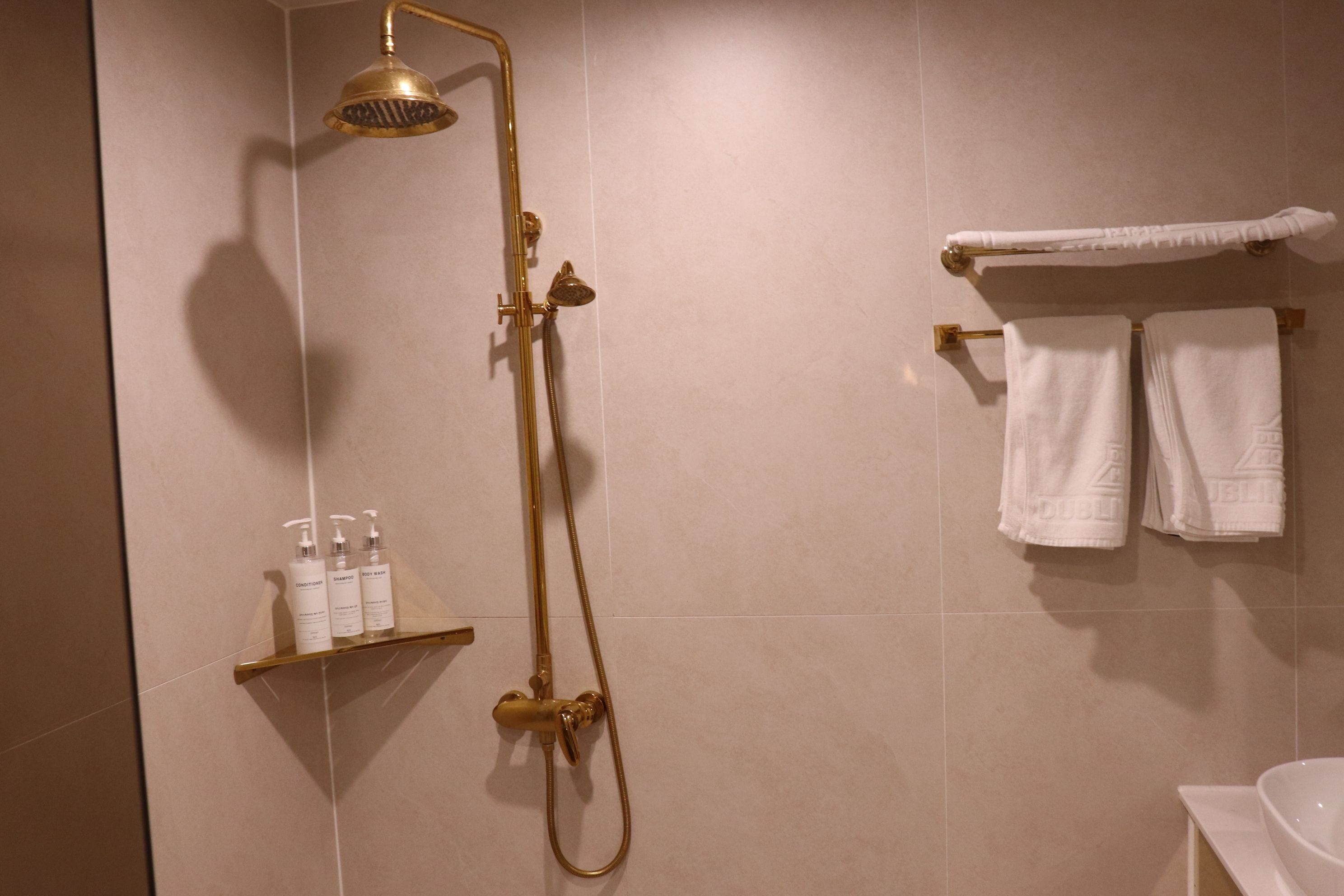 Kylpyhuone, jossa on kultainen suihku, valkoiset pyyhkeet ja hygieniatuotteet hyllyllä.