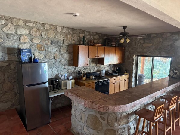A kitchen with stone walls, wooden cabinets, a built-in island with bar stools, a refrigerator, and a stove.