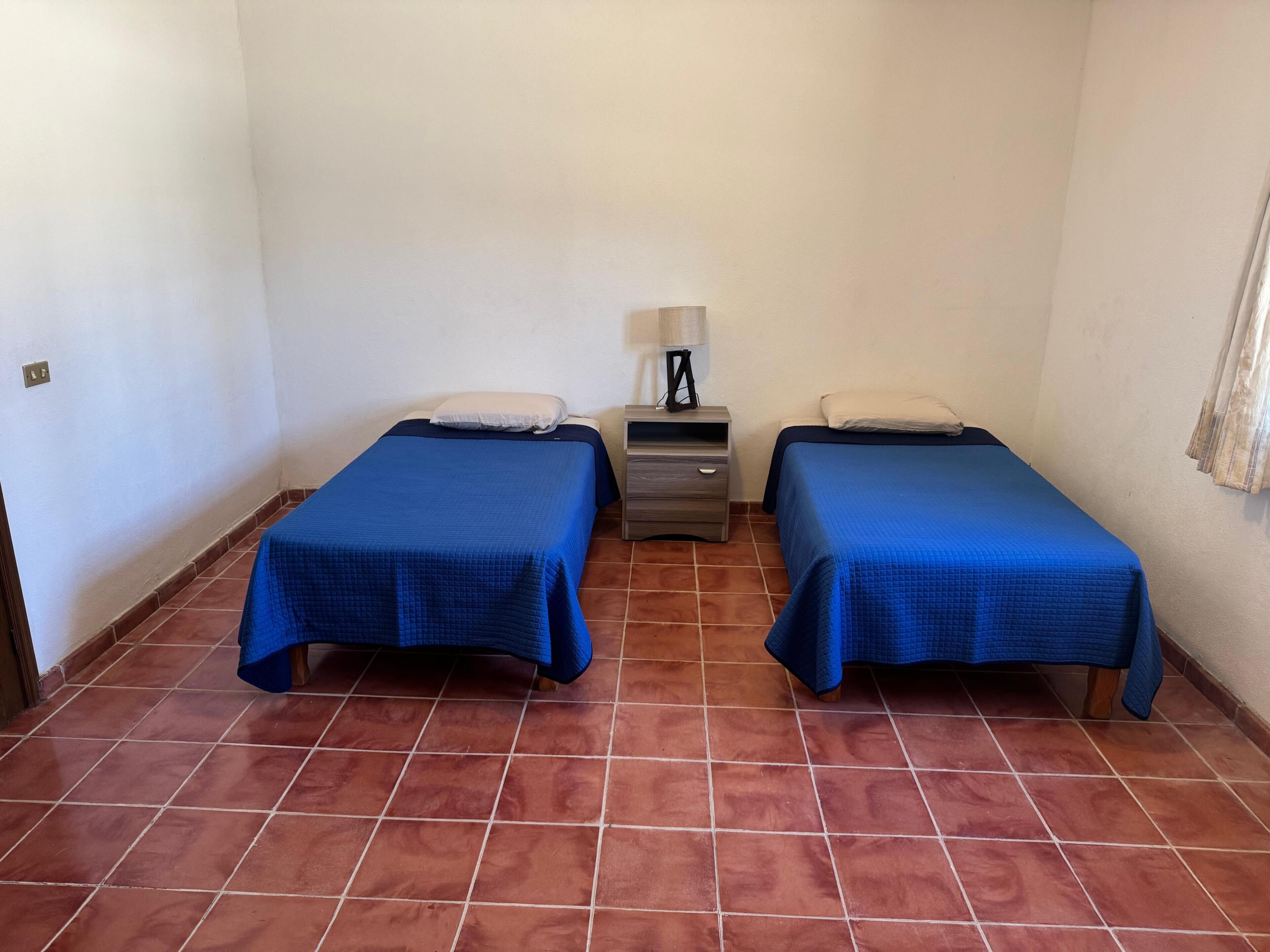 A room with two beds, a nightstand with a lamp, and a tiled floor.
