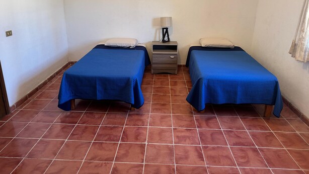A room with two beds, a nightstand with a lamp, and a tiled floor.