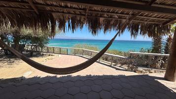 A hammock under a thatched roof, overlooking a coastal view with a clear sky.