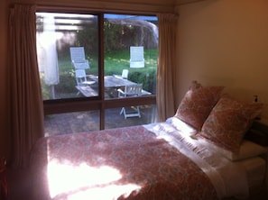 A bedroom with a large window, a bed with patterned bedding, and a view of an outdoor patio with chairs and a table. - COSY PORTSEA COTTAGE (Melbourne)