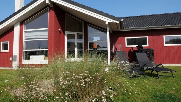 Un edificio rojo con un amplio ventanal y una chimenea. Hay sillas exteriores y un área verde con flores.