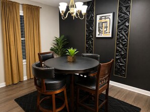 Un coin repas comprenant une table ronde, quatre chaises, un lustre, une plante en pot et un motif géométrique sur le mur.