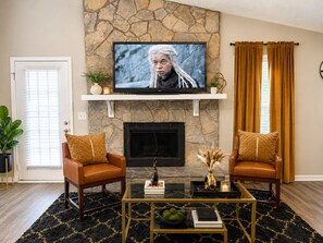 Ein Wohnzimmer mit Kamin aus Stein, einem darüber montierten Fernseher, zwei Ledersesseln, einem Couchtisch und einem schwarz-goldenen Teppich.