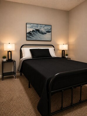 Une chambre avec un lit en métal noir, deux tables de chevet avec des lampes, un grand tableau encadré au mur et un tapis beige.