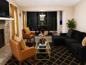 Un salon doté d’une cheminée, de fauteuils en cuir, d’un canapé noir, d’une table basse en verre et d’un coin repas agrémenté d’un lustre.