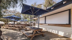 Un patio al aire libre con mesas de picnic de madera y sombrillas azules, un edificio blanco con puerta de garaje y árboles al fondo.