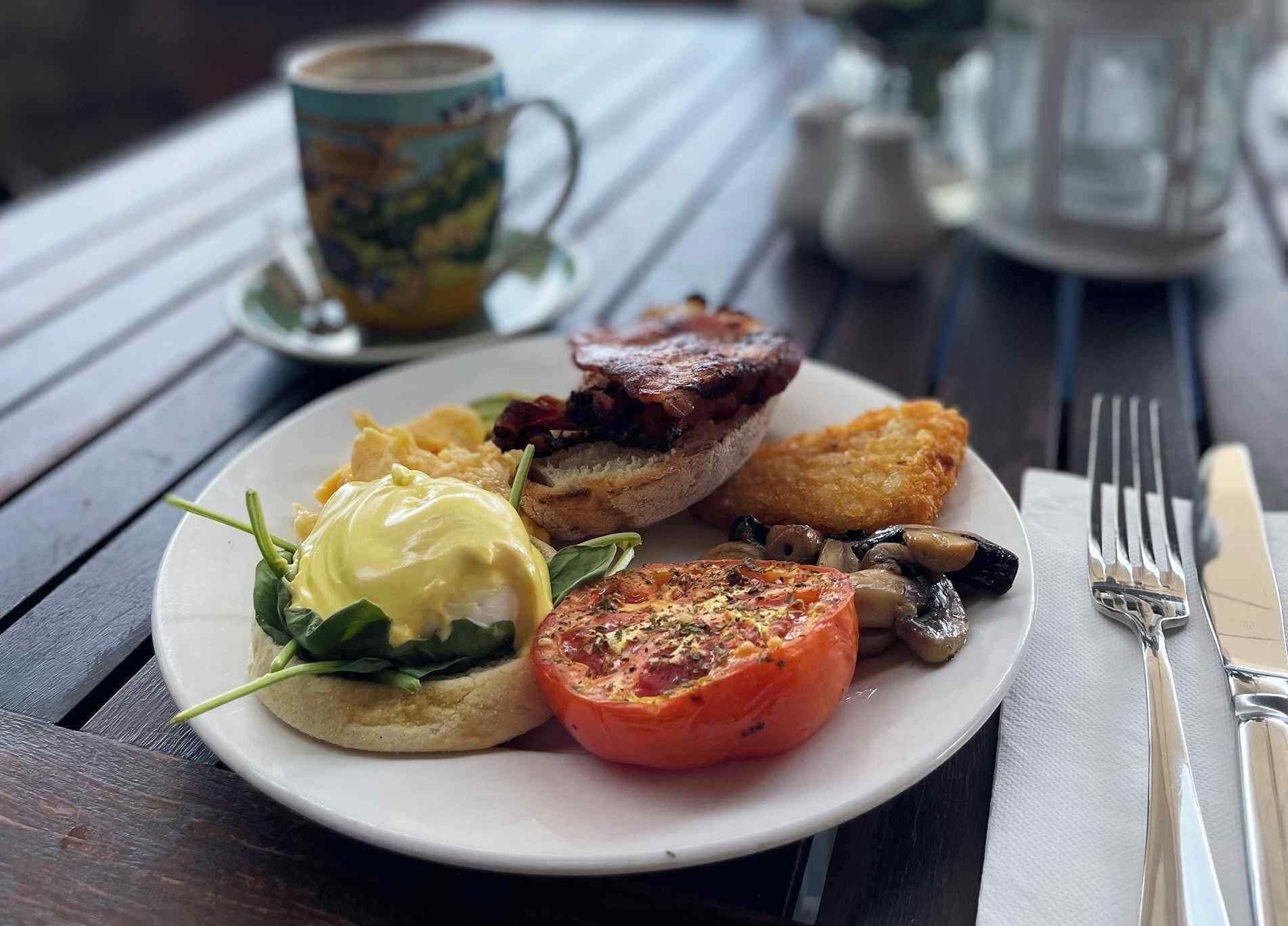 Un plato con huevos benedictinos, tomate a la parrilla, champiñones y un trozo de tocino sobre una mesa de madera.