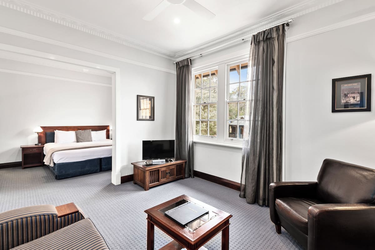 A hotel room with a bed, a TV on a wooden stand, a leather armchair, a small table, and a window with curtains.