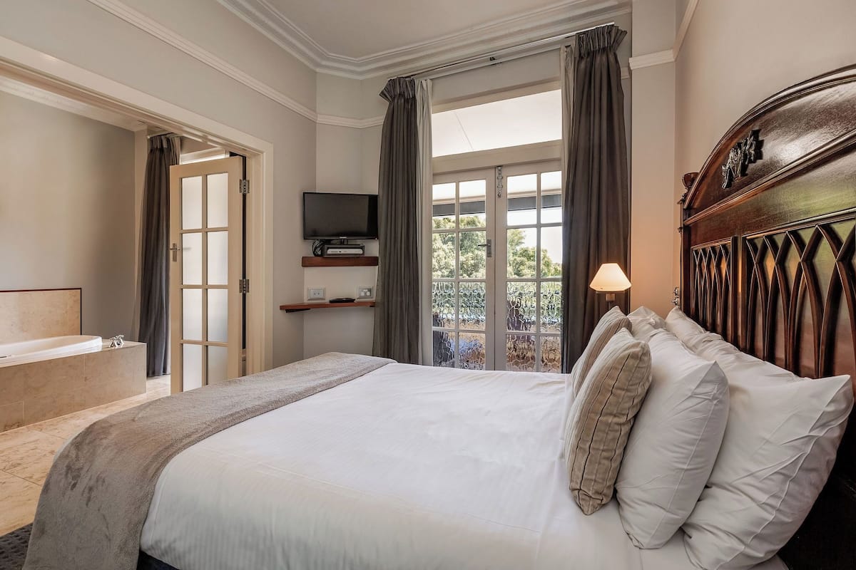 A bedroom with a large bed, a television, a balcony with curtains, and a bathtub.
