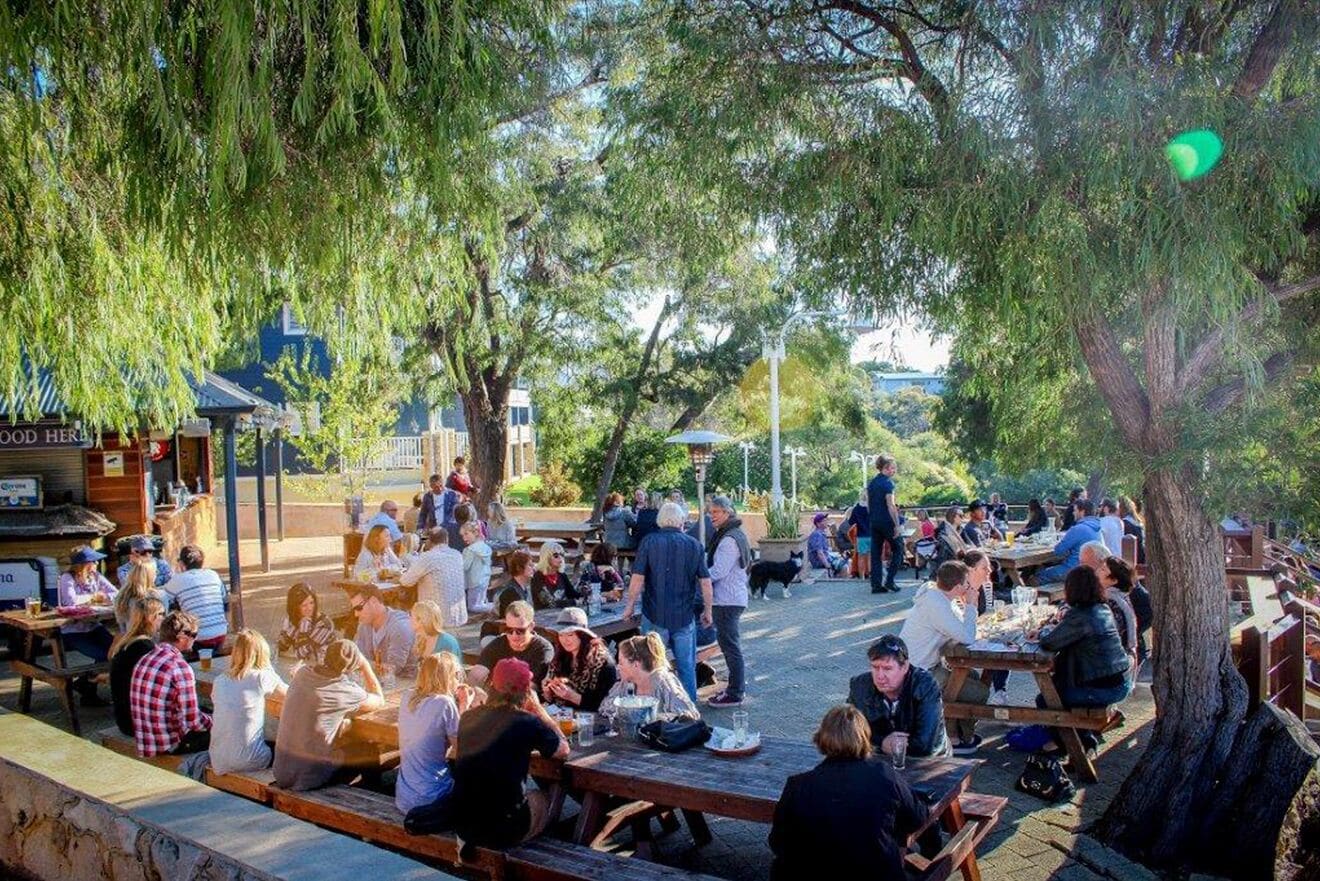 An outdoor dining area with wooden tables and benches, surrounded by trees.
