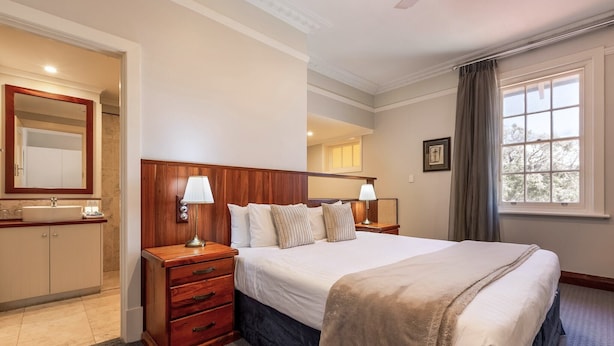 Habitación de hotel con una cama grande, lámparas de noche, una mesita de noche de madera, cómoda y un baño con espejo, además de una ventana con cortinas.