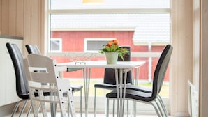 Uma área de refeições com mesa e cadeiras, uma planta em vaso com flores, e vista para um edifício vermelho lá fora.