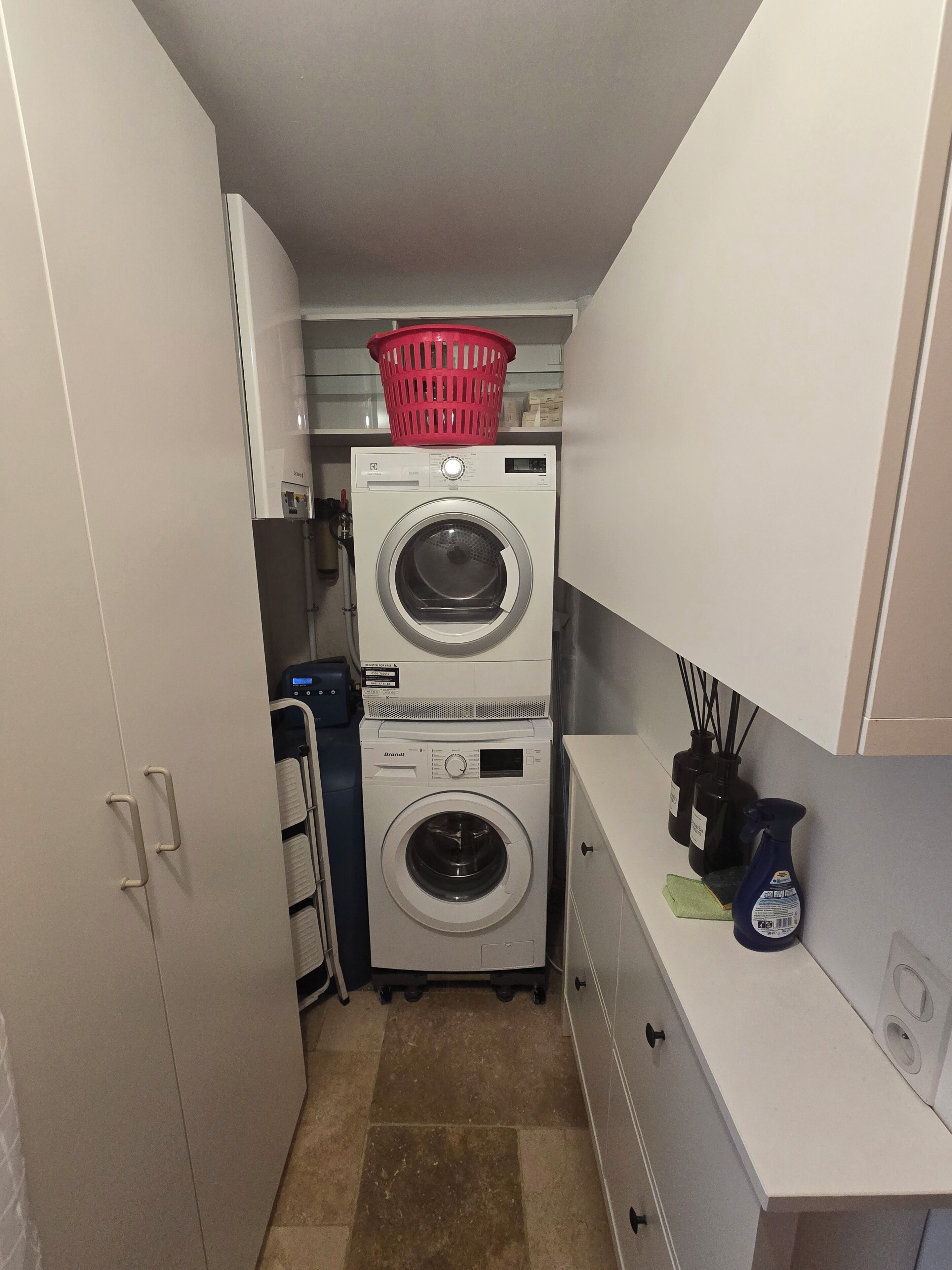 Une buanderie équipée de deux machines à laver superposées, un panier à linge rouge posé sur le dessus, et un meuble blanc sur lequel se trouve un vaporisateur.