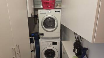 Une buanderie équipée de deux machines à laver superposées, un panier à linge rouge posé sur le dessus, et un meuble blanc sur lequel se trouve un vaporisateur.