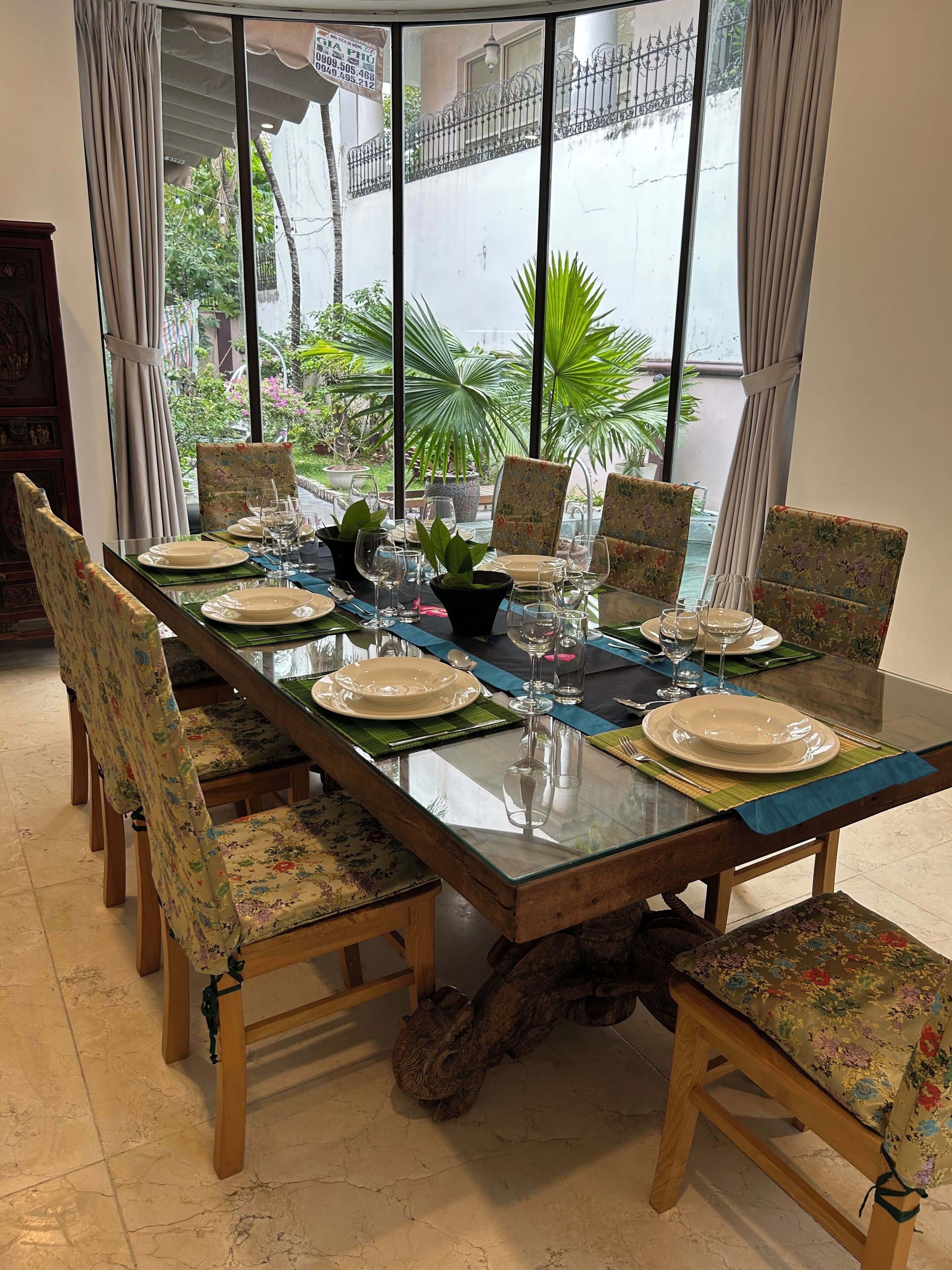 Mesa de comedor lista para seis personas con sillas de estampado floral, una mesa de vidrio y vistas a follaje exterior.