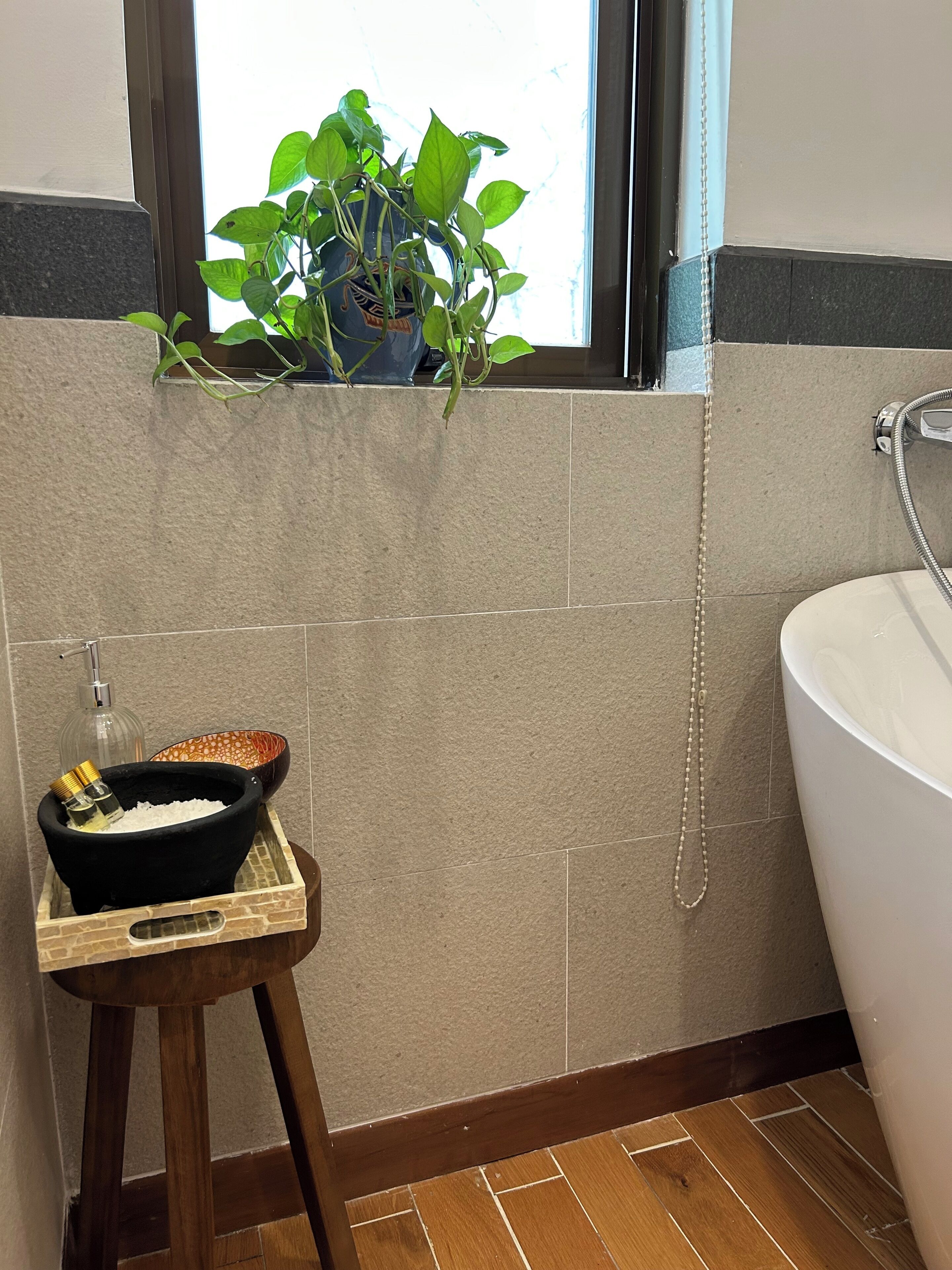Een badkamer met een bad, een douche, een raam met een plant en een klein krukje met een kom en flessen.