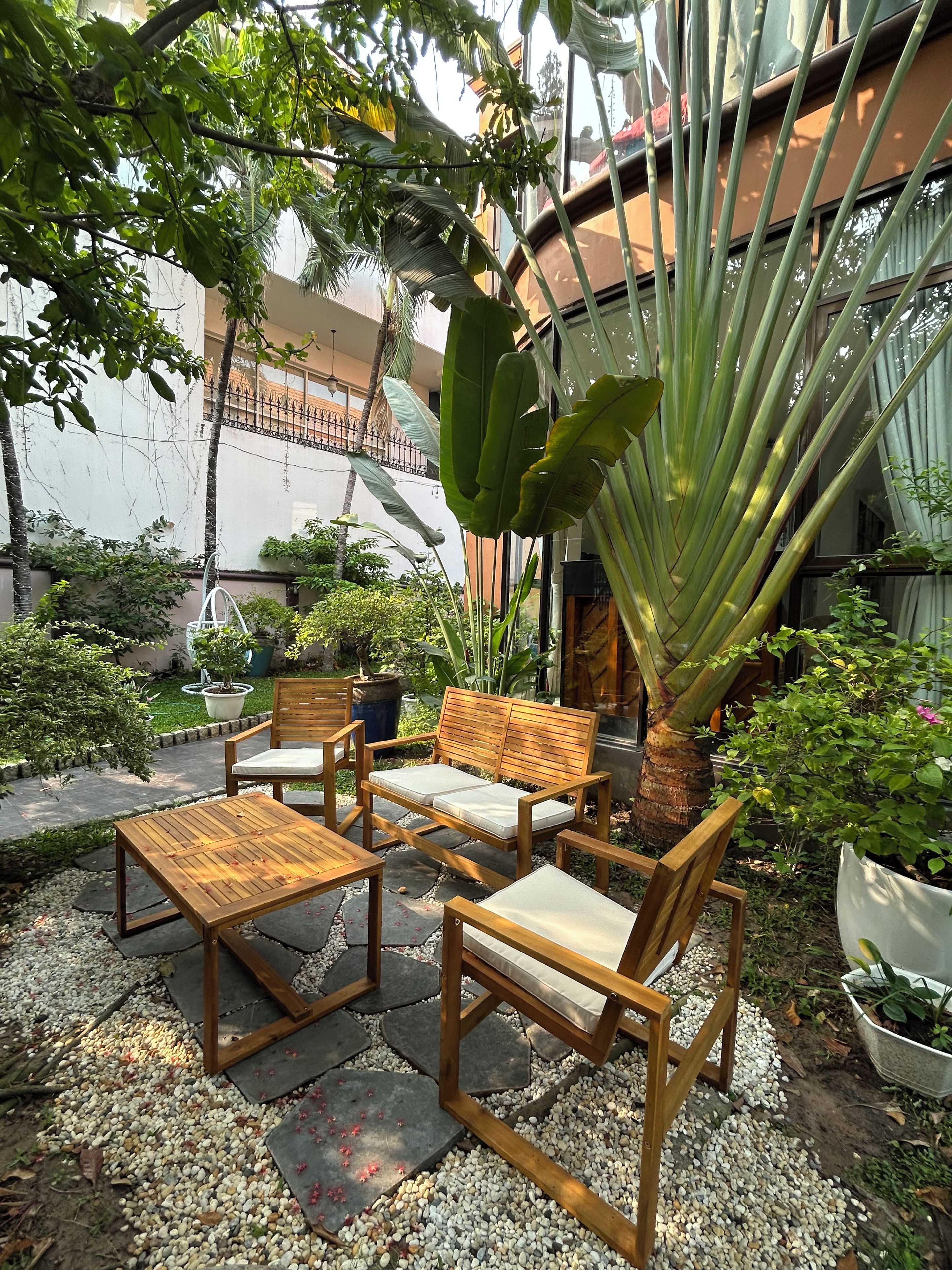 Área de pátio com um banco e mesa de madeira, cercada por vasos de plantas e uma bananeira grande.