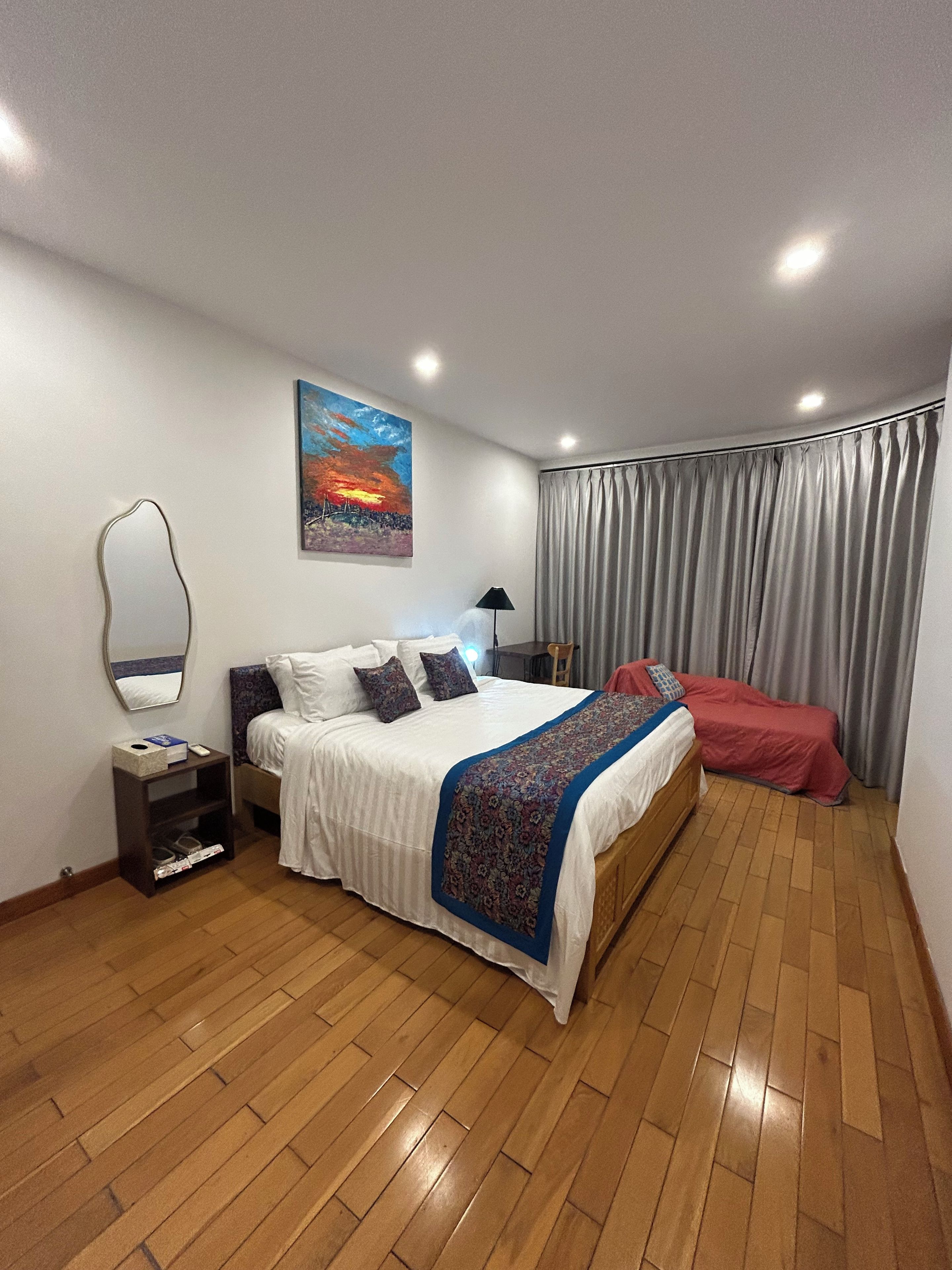 A bedroom with a bed, a nightstand, a lamp, a painting, and a window with curtains.